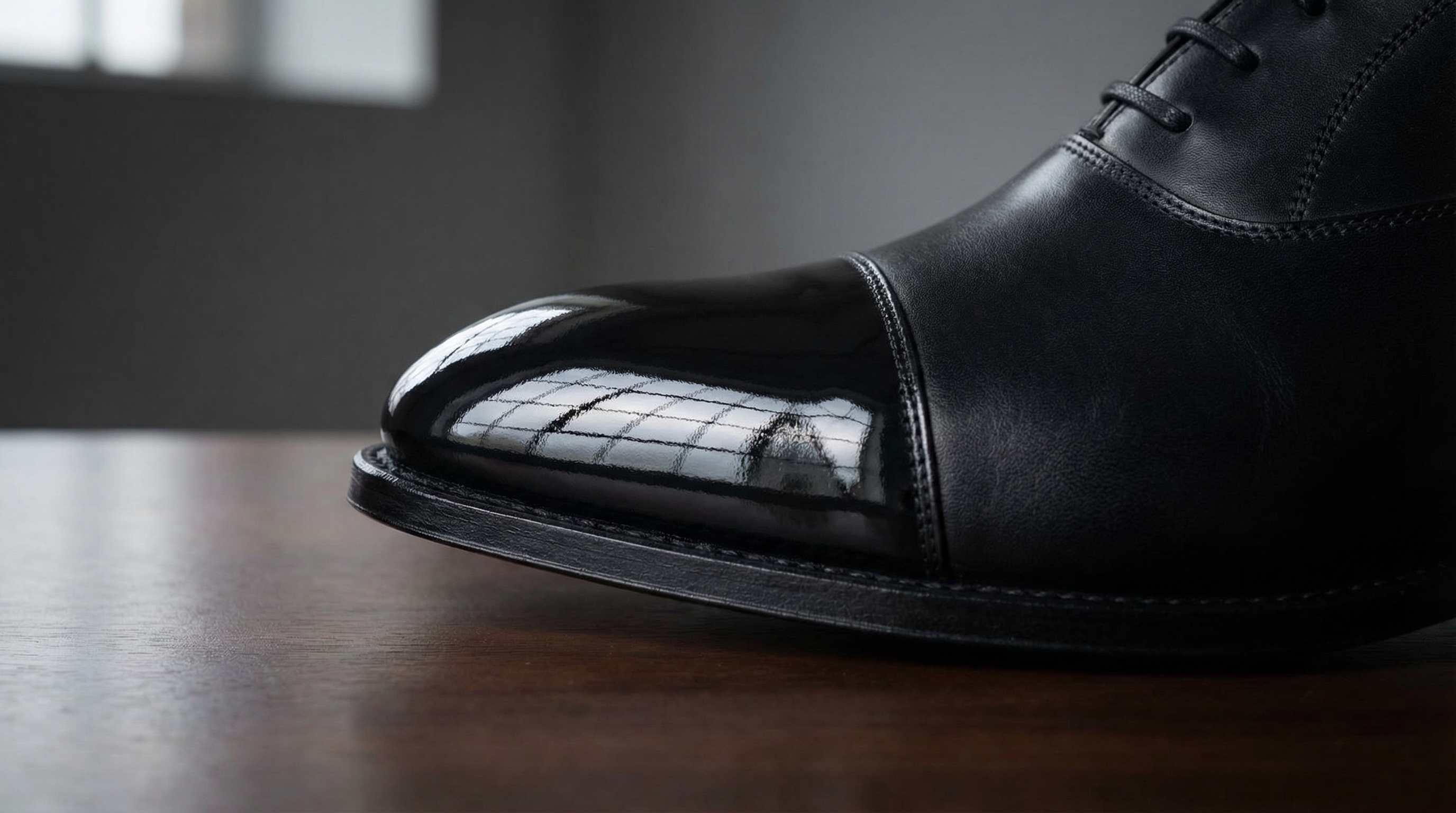 Commercial product photography, low-angle side view, focusing on the toe cap of a black calfskin shoe. The toe displays a perfect mirror-like reflection of a nearby window, showing the transition from a matte vamp to a high-gloss toe. Crisp, sharp lighting highlighting the reflection and leather grain.