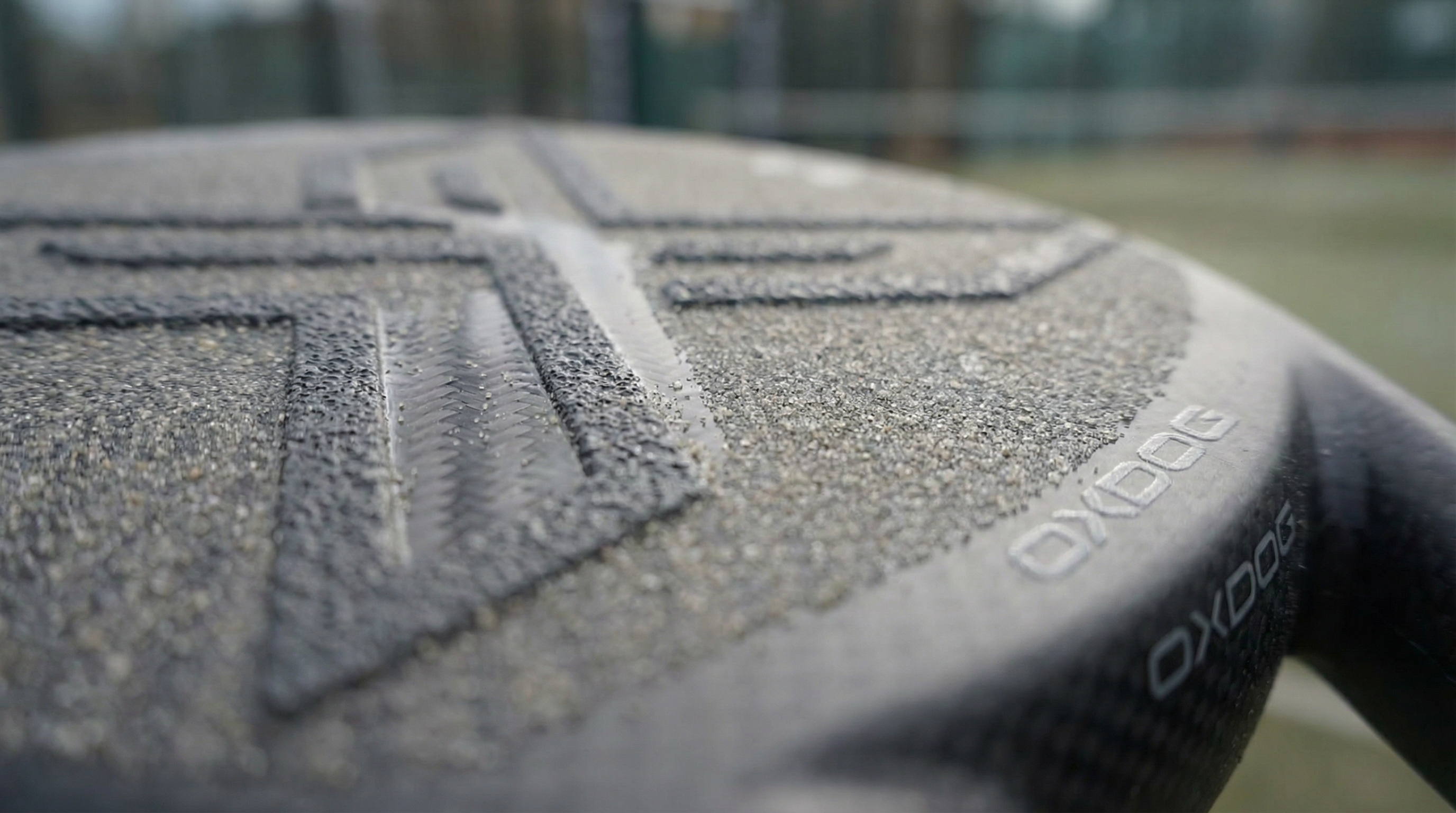 Photorealistic close-up image of the textured sandy surface of an oxdog padel racket, showing the intricate grip patterns and the 8K carbon weave underneath.