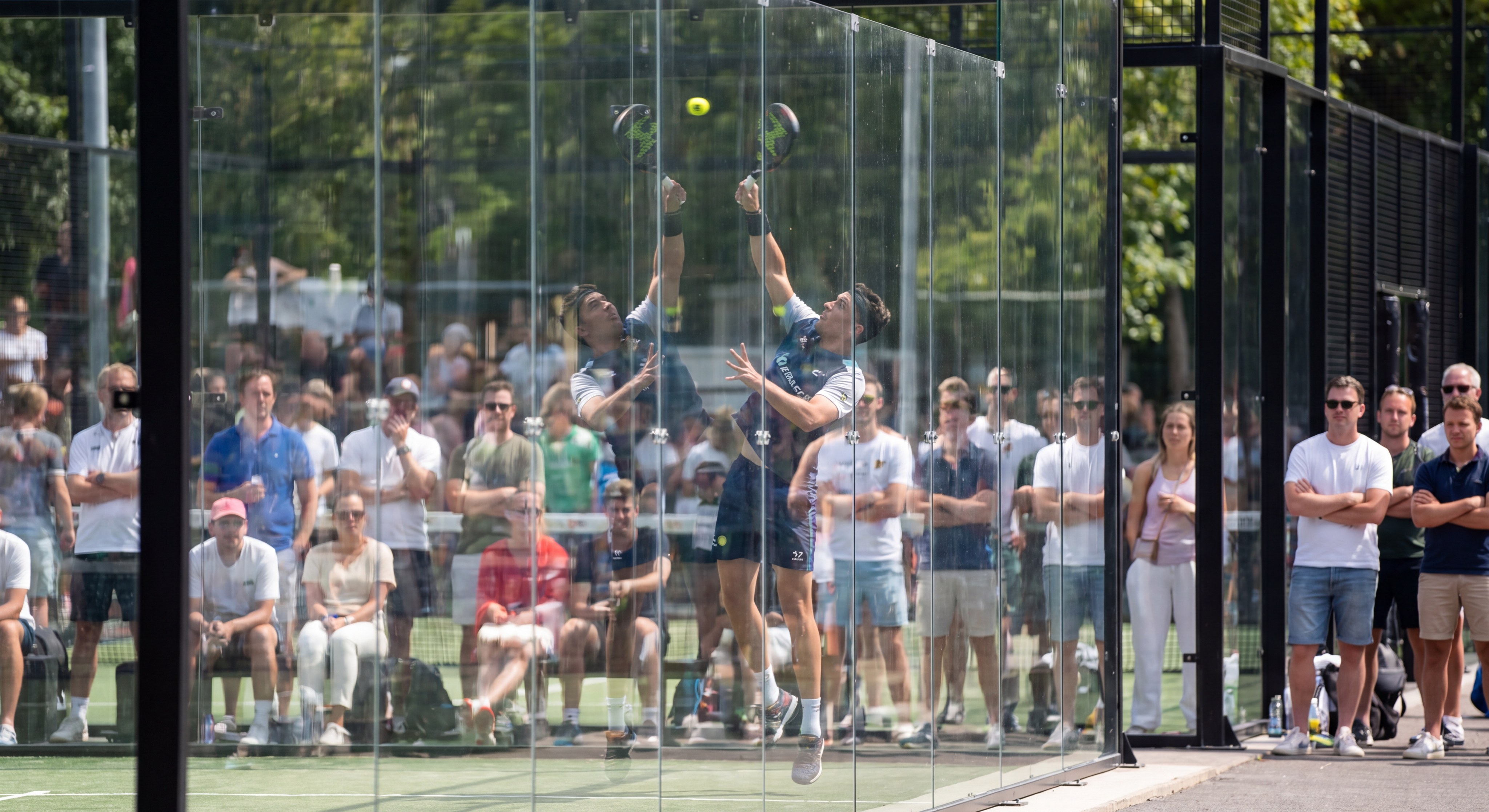 An action shot of players seen through the crystal-clear glass walls of a padel court
