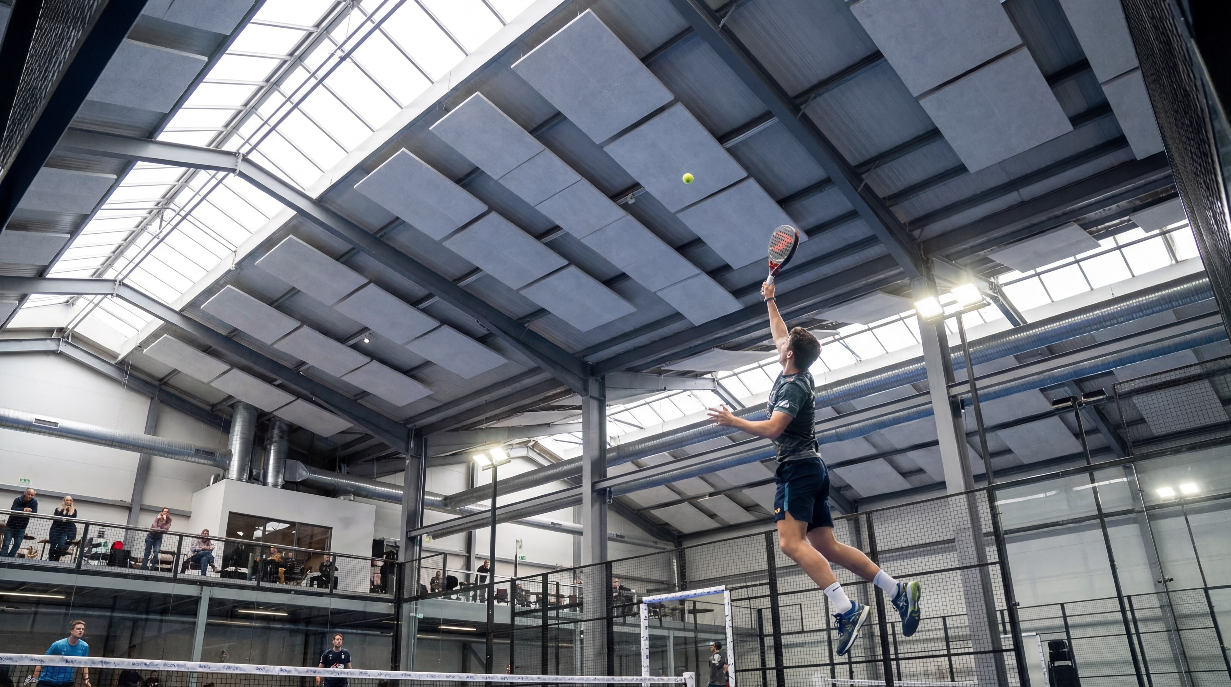 An indoor padel facility with ultra-high ceilings and a player hitting a lob