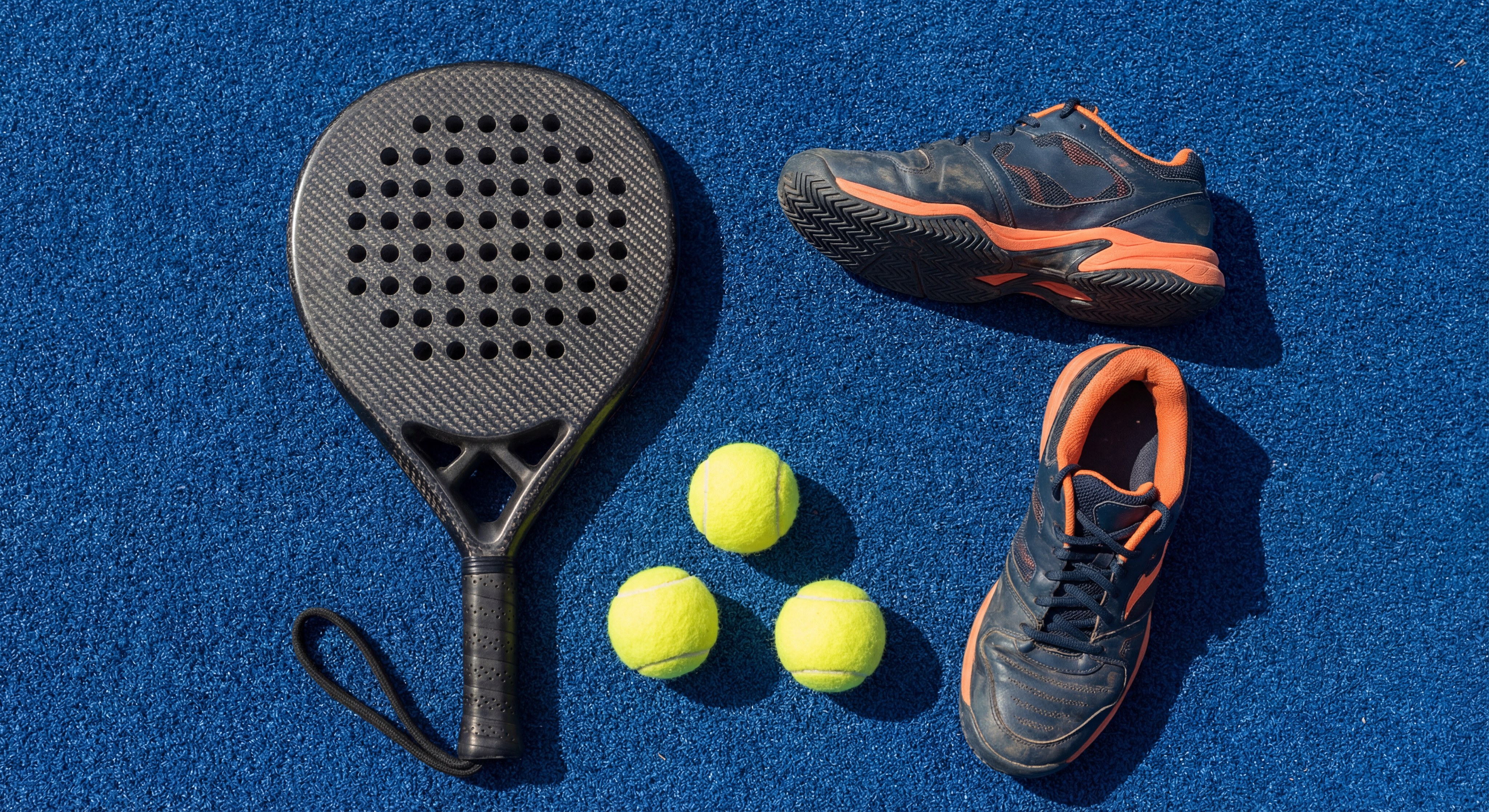 Photorealistic arrangement of a carbon fiber racket, specialized shoes, and three bright yellow padel balls on a textured blue turf.