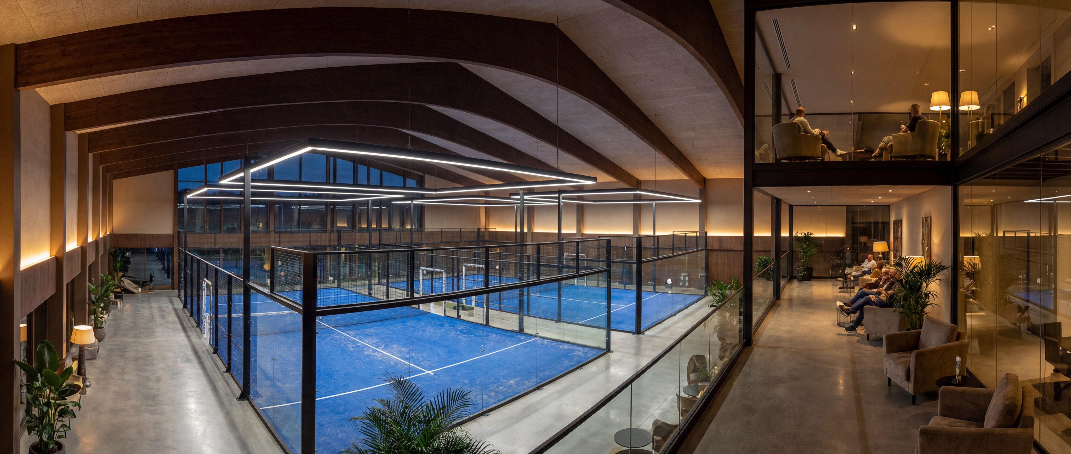 A stunning wide view of a luxury indoor padel club with multiple courts, professional LED lighting, and a glass-walled viewing gallery.