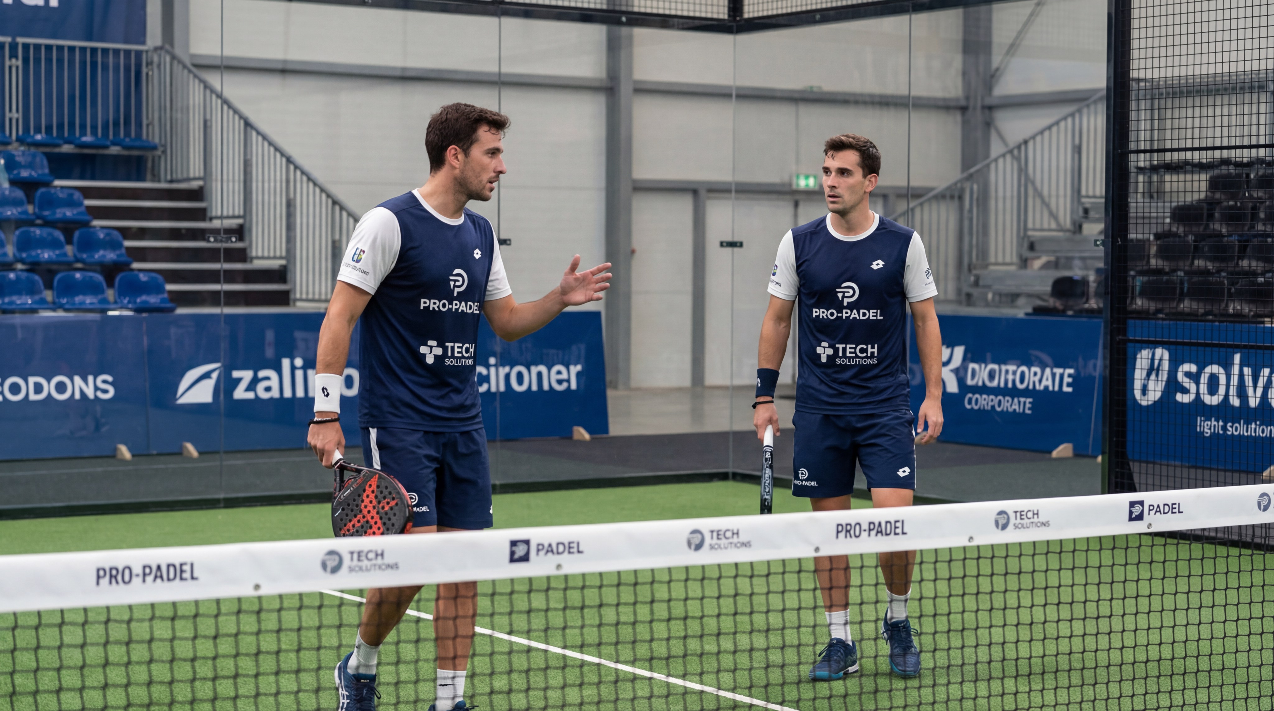 Two professional padel players communicating mid-point, one at the net and one at the baseline, showcasing strategic doubles positioning.