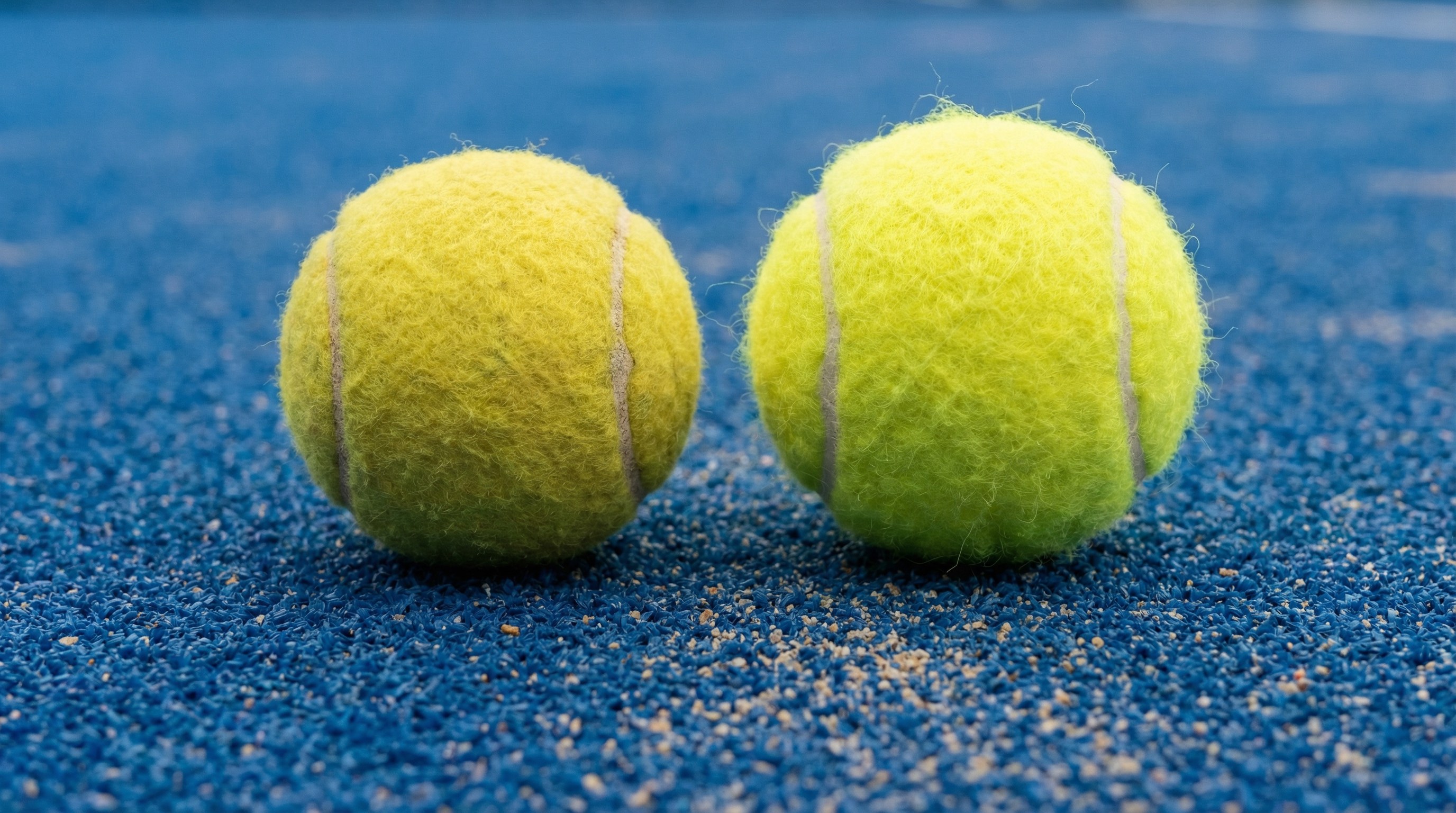 Photorealistic close-up of a padel ball and a tennis ball side-by-side on a blue court surface, showing slight differences in felt texture and size.