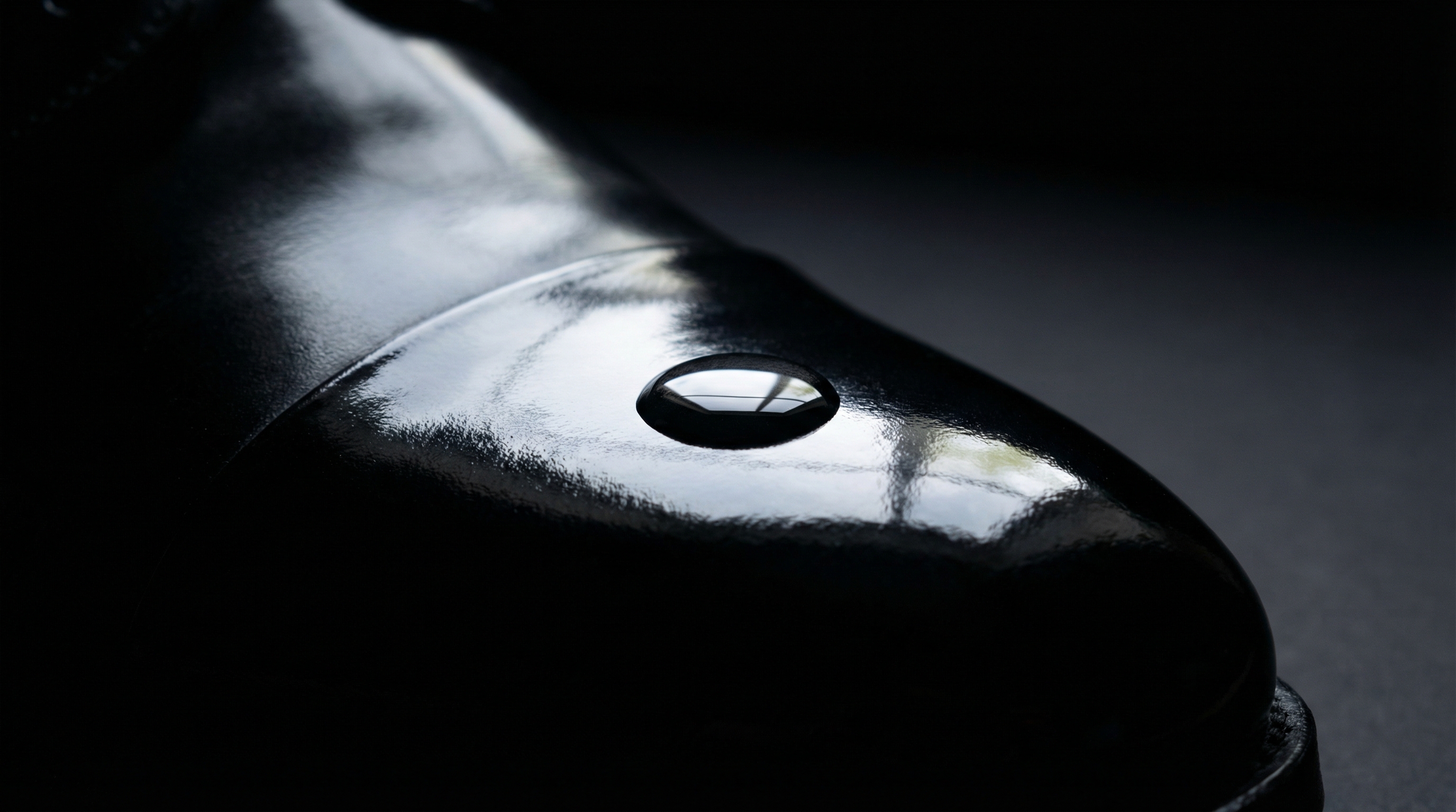 Macro shot of a single water droplet on a glass-like, mirror-polished black leather shoe toe cap.